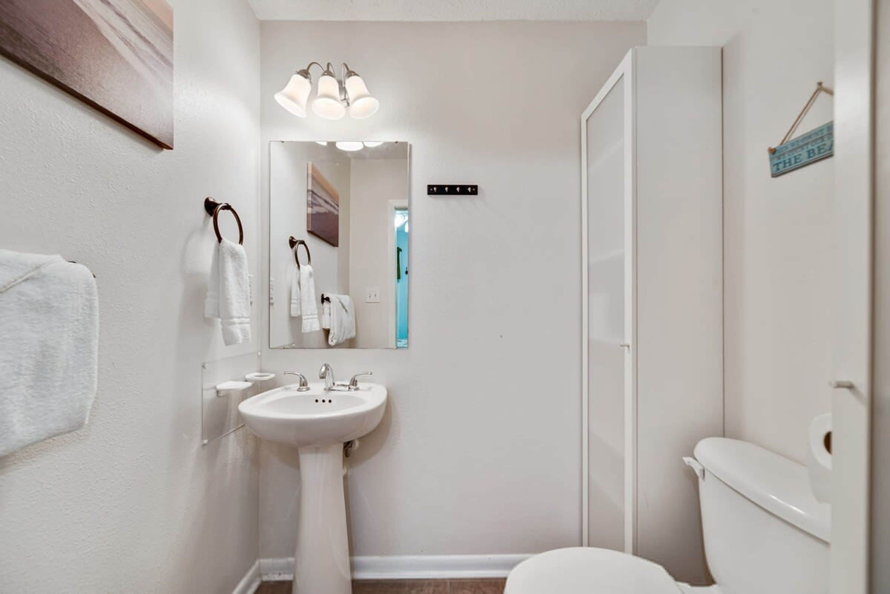 Een badkamer met een wit wastafelmeubel, spiegel en toilet. Er is een handdoekrek met handdoeken en een bordje aan de muur.