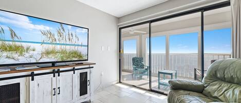 A modern living room with a large flat-screen TV, a leather sofa, and a sliding glass door leading to a balcony with a view of the sky and clouds.