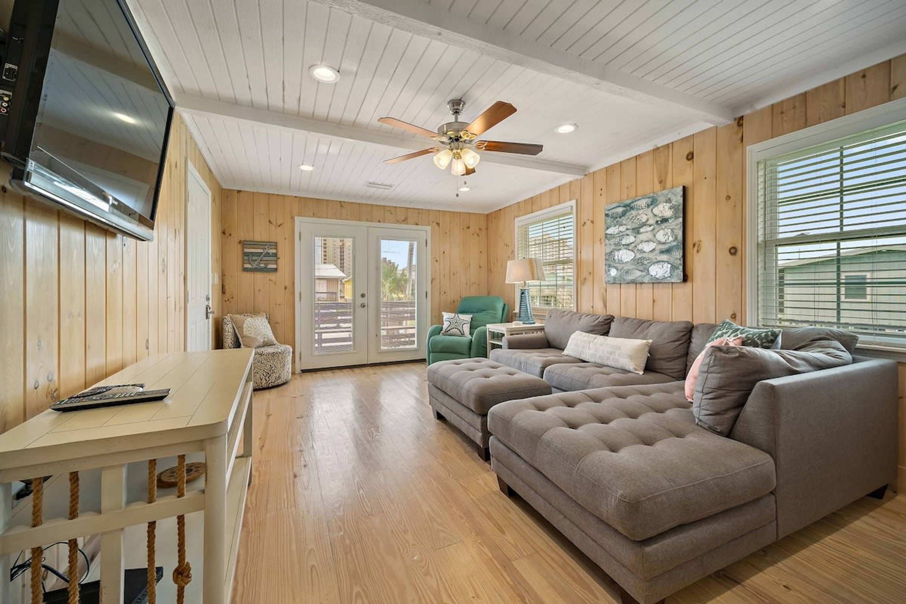 A cozy living room with a wooden interior, a ceiling fan, a television, and a large window with blinds.