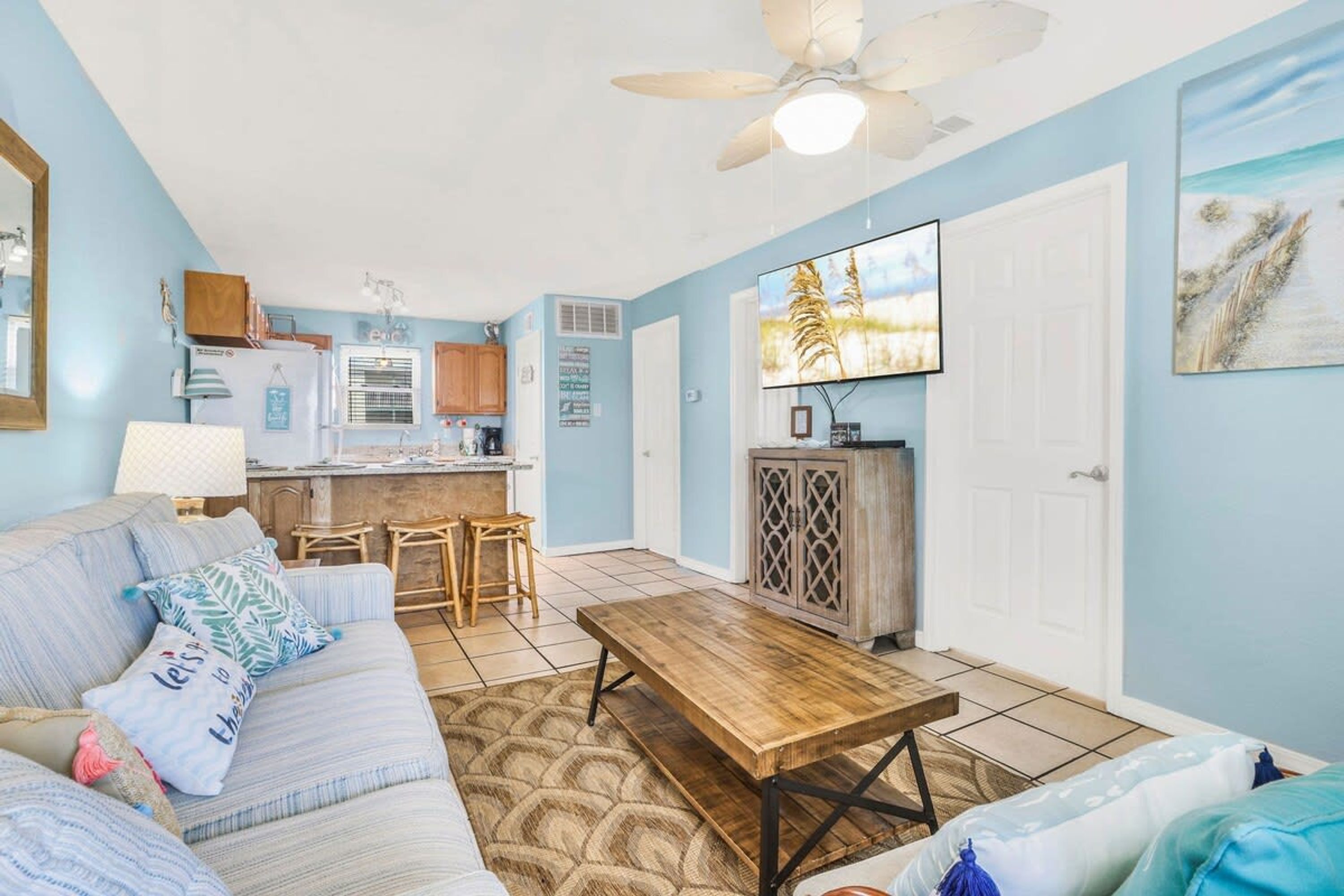 A living room with a sofa, coffee table, and a TV. There is a kitchen area with a refrigerator and cabinets.