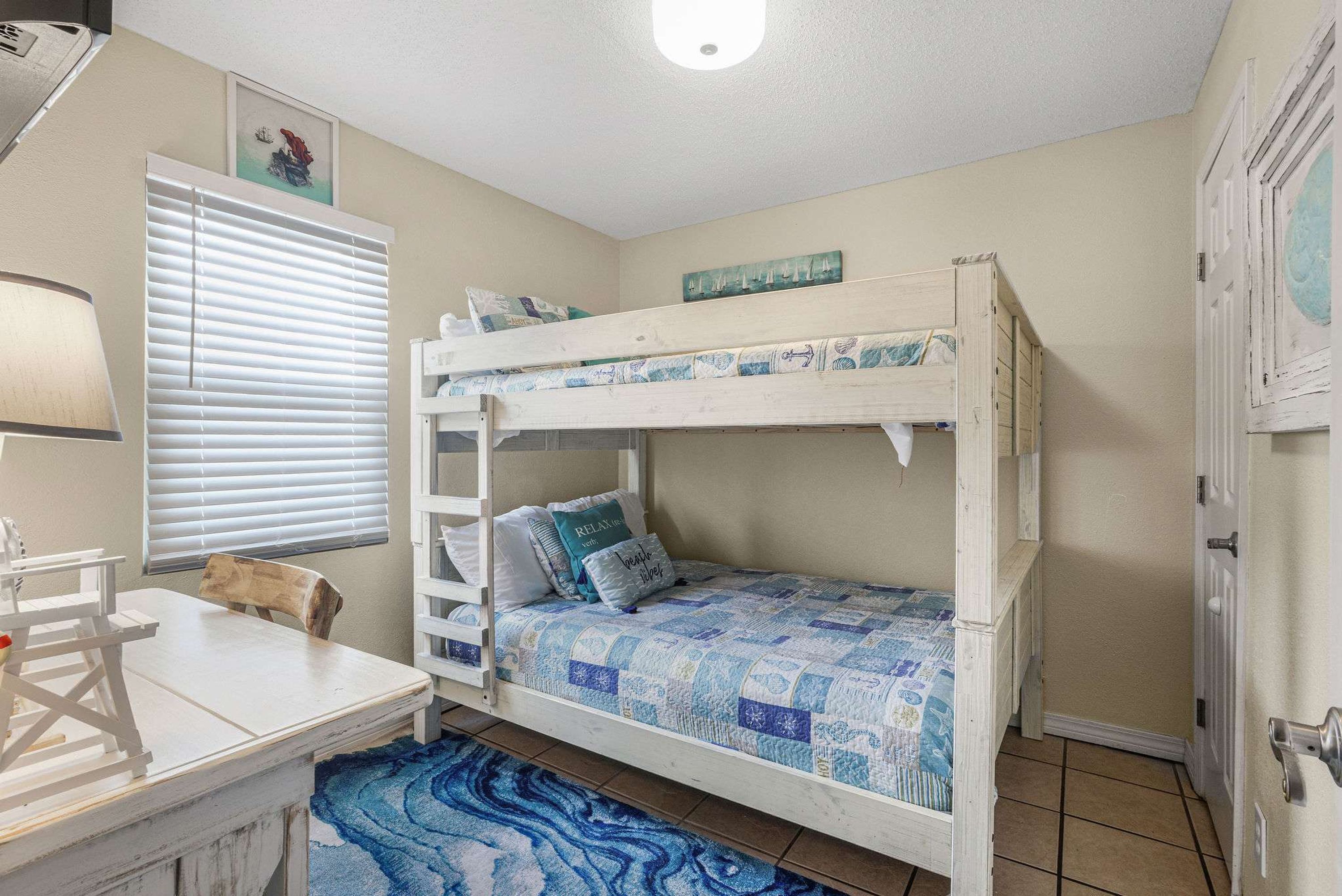 A bunk bed with a blue quilt, a desk with a lamp, and a window with blinds.