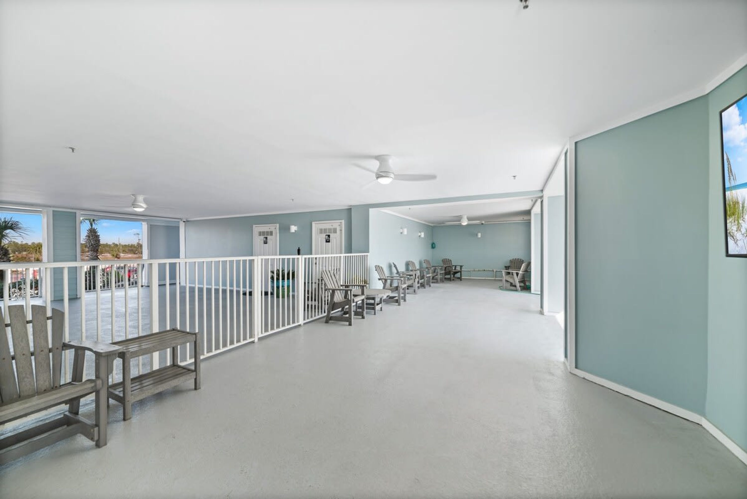 A spacious indoor area with a row of wooden chairs and a bench along a white railing, a ceiling fan, and a view through large windows.