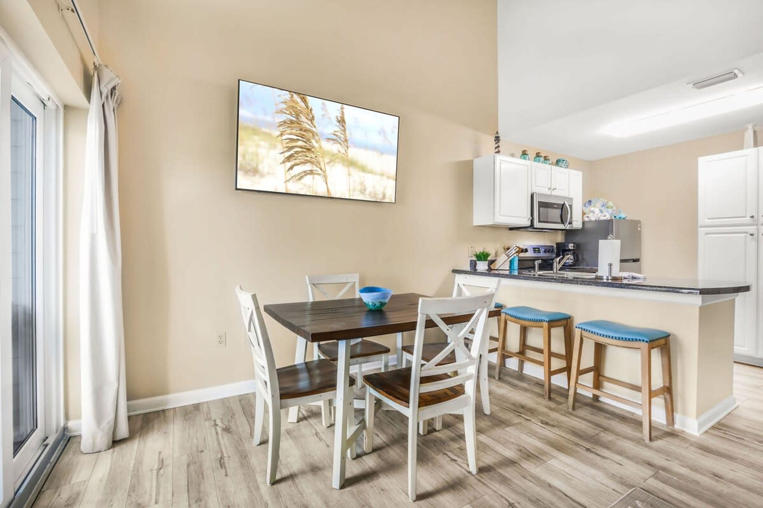 Una cocina moderna con armarios blancos, un microondas y un fregadero. Hay un comedor con una mesa de madera y sillas blancas.