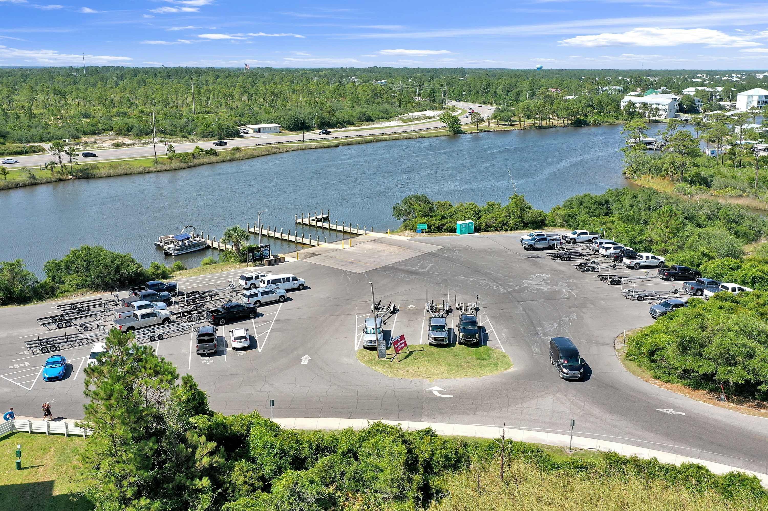 A parking lot adjacent to a body of water with boats docked, a road leading to a bridge, and a small dock.