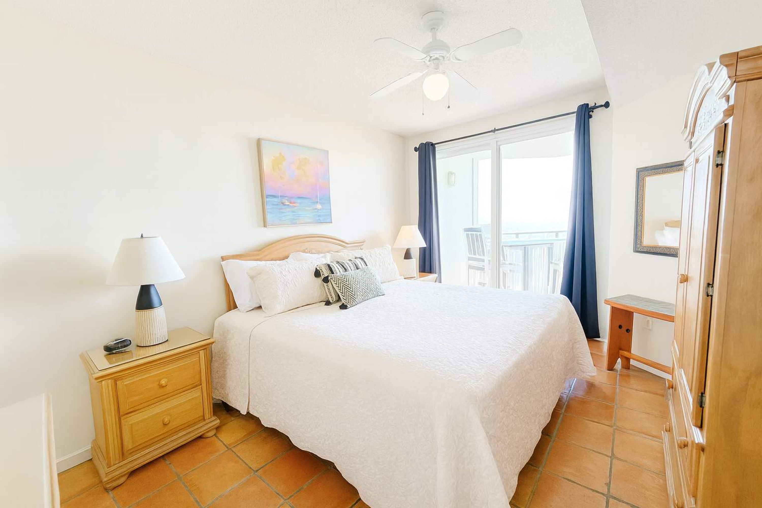 Une chambre à coucher comprenant un lit, une table de chevet, une lampe, un ventilateur de plafond et une vue sur l’extérieur.