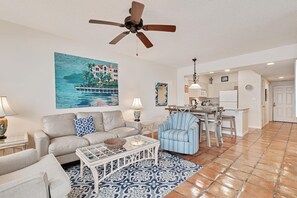 A living room with a sofa, coffee table, and patterned rug. There is a dining area with a table and chairs. A ceiling fan is visible. - Silver Beach 302 Beachfront 3rd Floor Pool (Orange Beach)