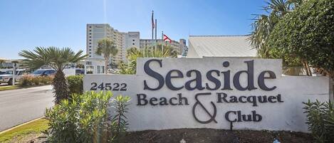 A sign for Seaside Beach & Racquet Club with a clear view of the surrounding area including a road and buildings.