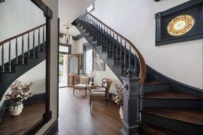 A grand staircase with a dark wooden finish and black balusters, leading to a room with a large stained glass window and a view of the outdoors. - Historic Restoration | Whitefield Square (Savannah)