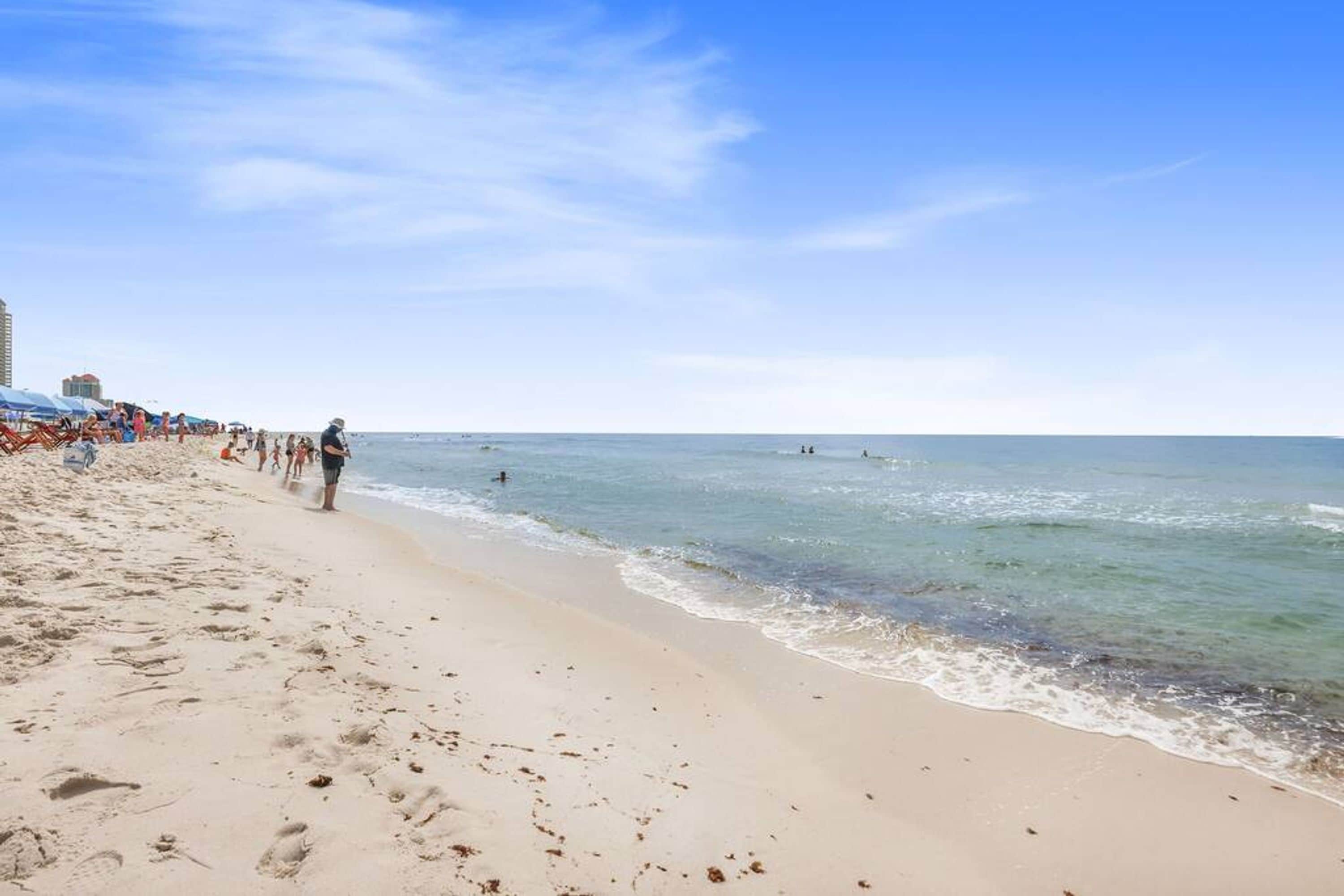Une plage avec un ciel dégagé, des gens qui se promènent le long du rivage et quelques parasols.