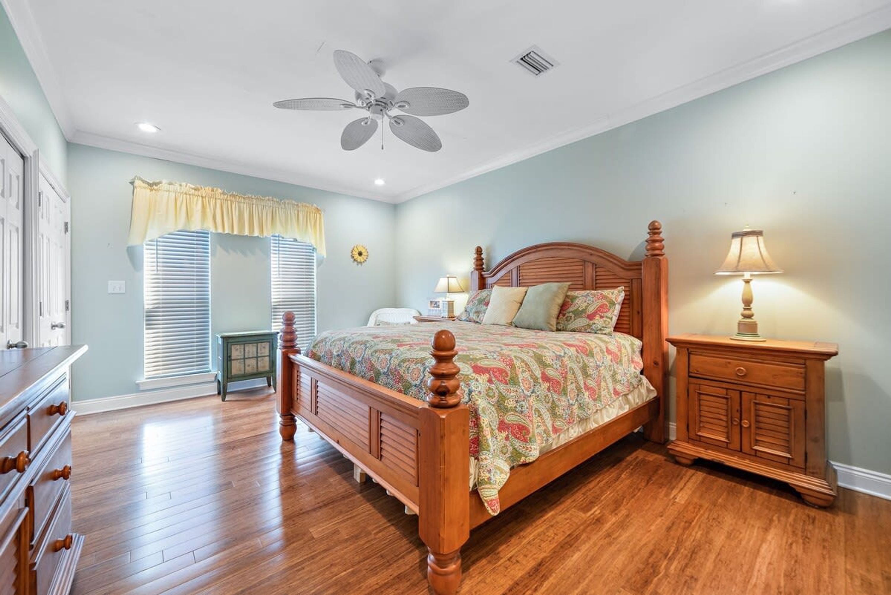 A bedroom with a wooden bed, a ceiling fan, a nightstand with a lamp, and a window with blinds.