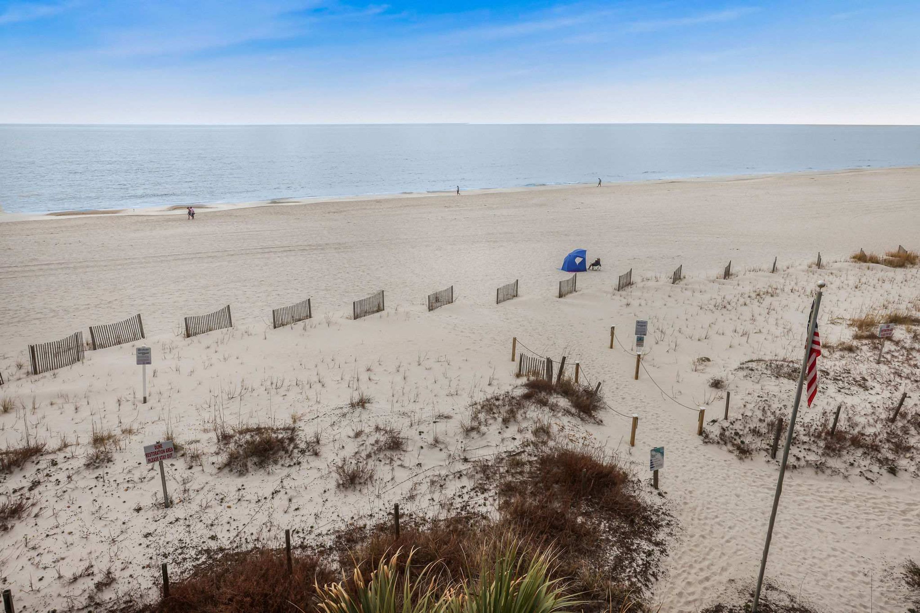 A beach with a sandy path leading to the water, a blue umbrella, and a flag.