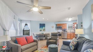 A living room with a sofa, ottoman, and armchair, a dining area with chairs, and a kitchen with appliances. - Sandpiper 1C- Stylish Gulf-side Affordable (Gulf Shores)