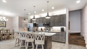 A modern kitchen with dark cabinets, a central island, pendant lighting, and a dining area with wooden chairs. - Hammock Dunes Beachside Cottage (Orange Beach)