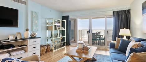 A modern living room with a sofa, coffee table, and a TV. There is a balcony with a view.