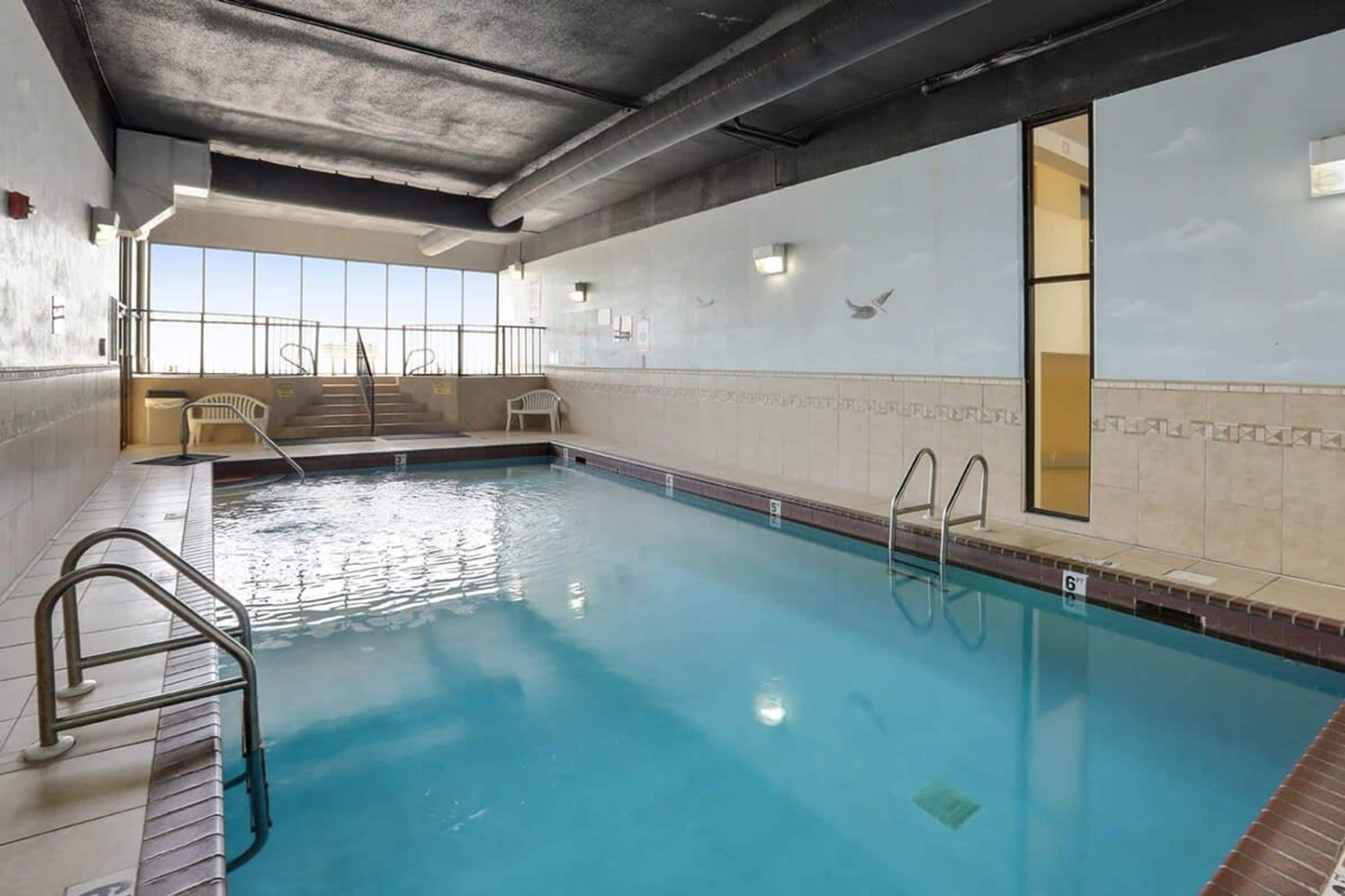 An indoor swimming pool with clear water, metal railings, and a view of the sky through large windows.