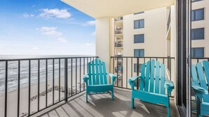 Ein Balkon mit zwei türkisfarbenen Adirondack-Stühlen, Blick auf das Meer und ein Gebäude mit zahlreichen Fenstern und Balkonen.