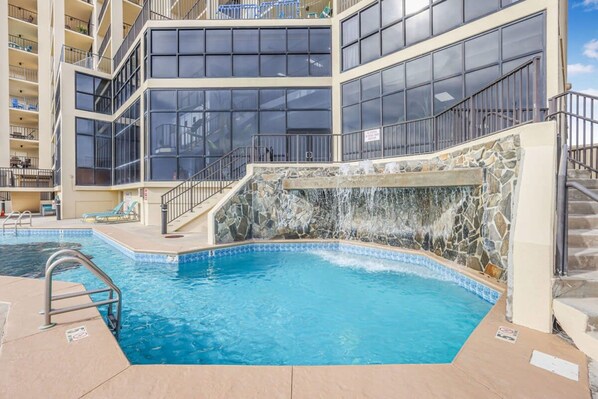 A swimming pool with a stone waterfall feature, surrounded by a multi-story building with balconies and large windows. - Phoenix II Orange Beach Beach Views Pools Fun (Orange Beach)