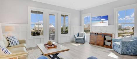 Un salón amplio con un sofá, un sillón y una mesa de centro. Hay ventanas grandes y un televisor montado en la pared.