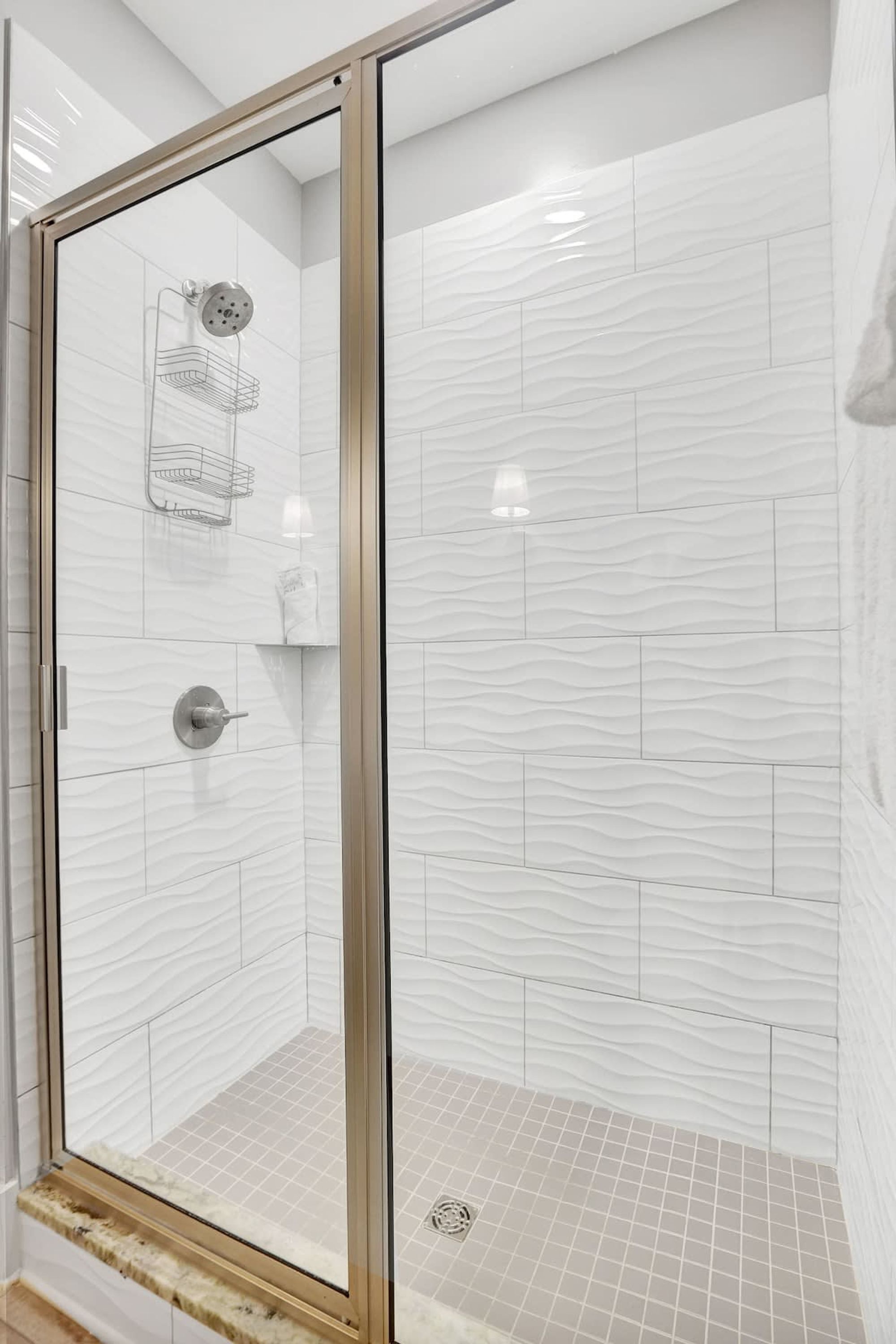 Ducha con cerramiento de vidrio, azulejos blancos con textura, un cabezal de ducha de mano y un perchero para toallas.