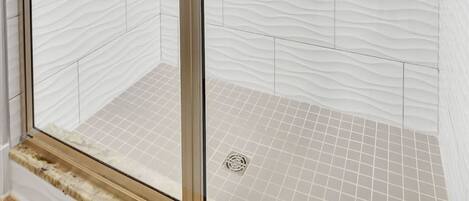 Ducha con cerramiento de vidrio, azulejos blancos con textura, un cabezal de ducha de mano y un perchero para toallas.