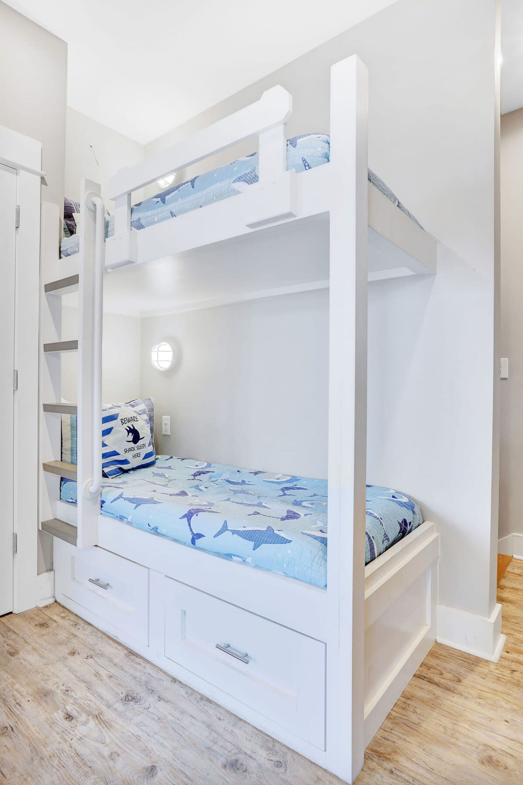 A bunk bed with a lower bunk and a pull-out desk, white walls, and a wooden floor.