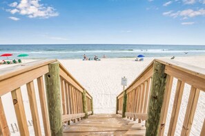 Un escalier en bois mène à une plage de sable où l’on aperçoit, au loin, des parasols et des gens.