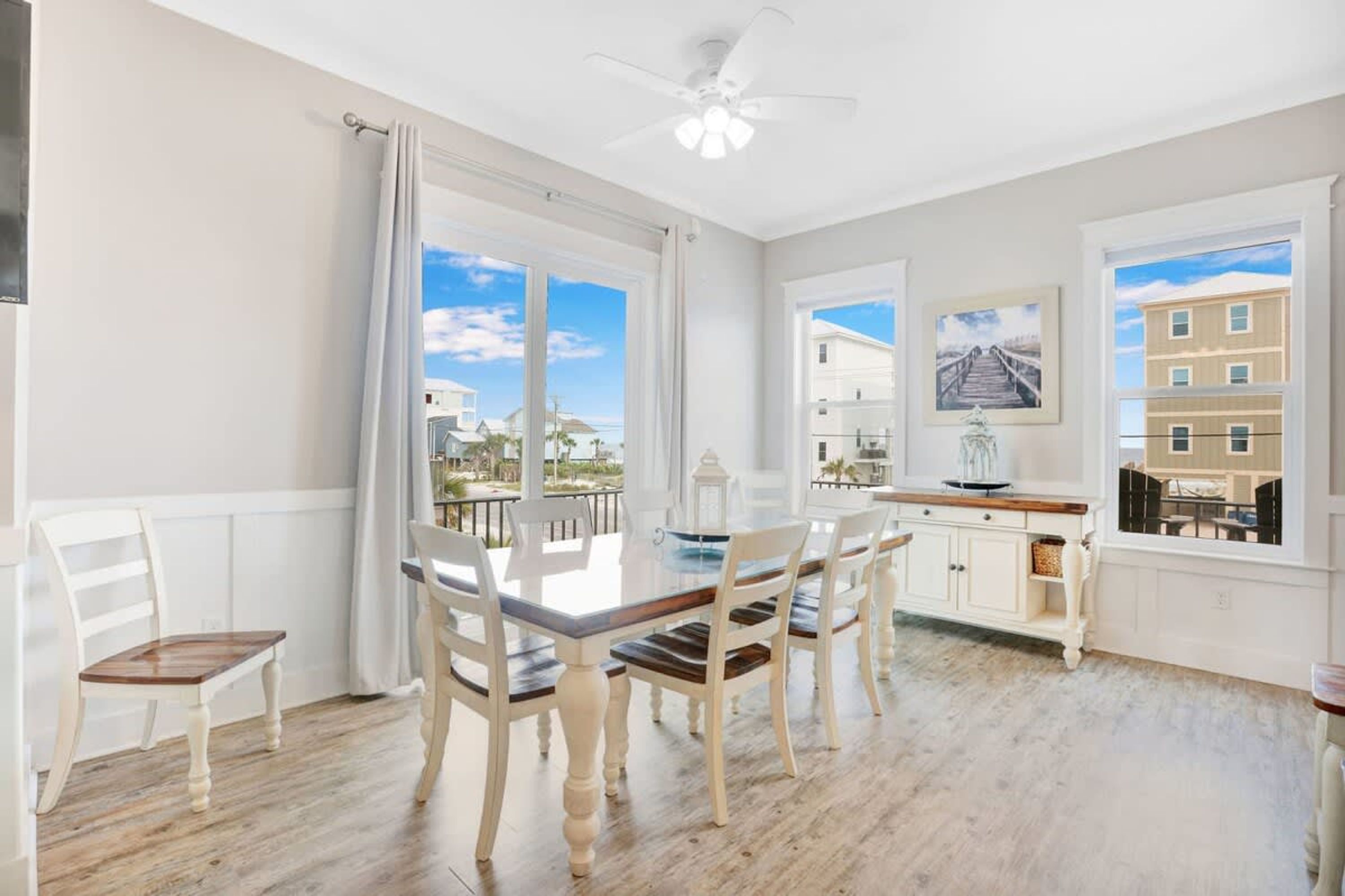 A dining area with a table and chairs, a ceiling fan, and large windows with a view of buildings and a beach.
