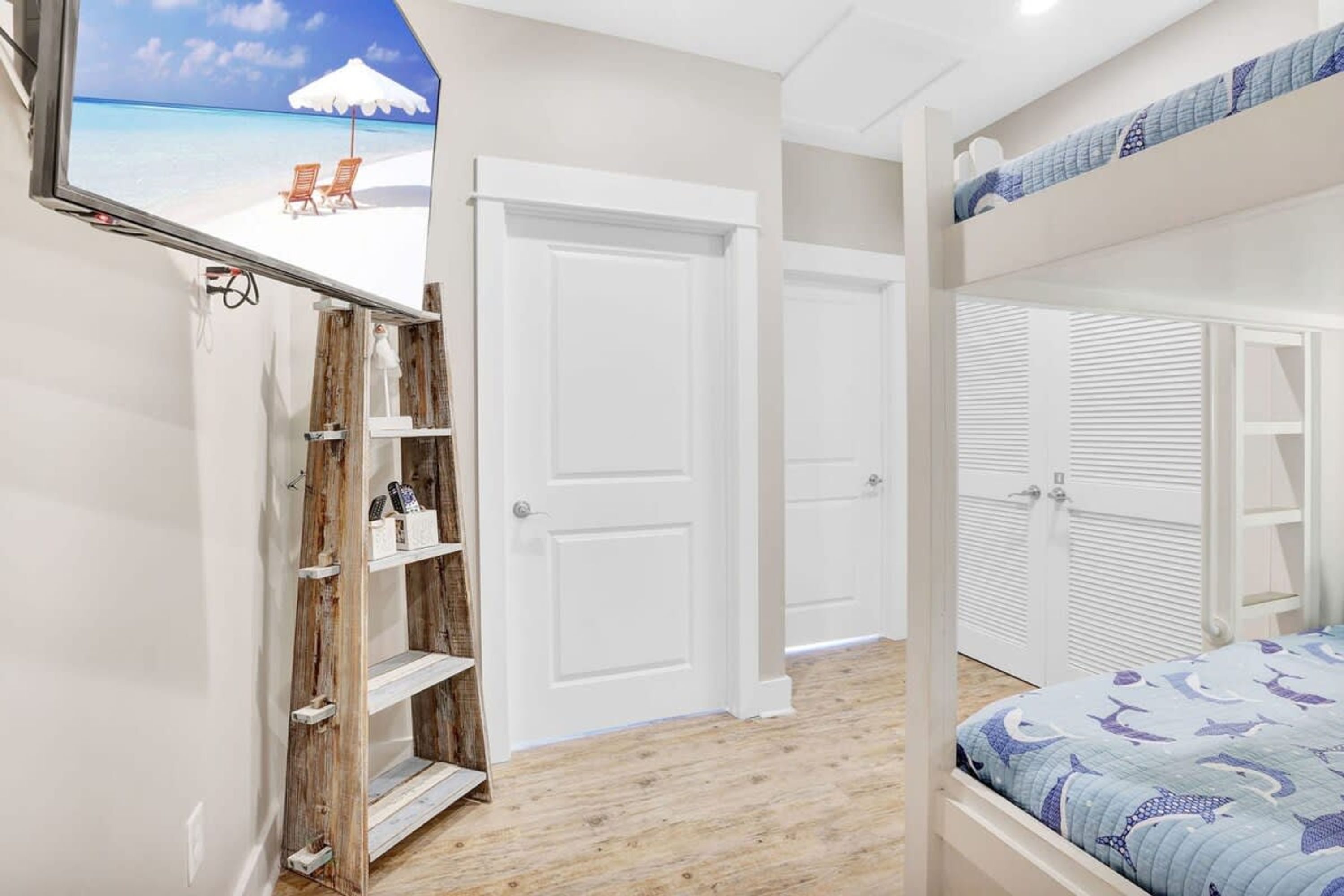 A bedroom with a bunk bed, a wooden shelf unit, a white door, and a wall-mounted TV displaying a beach scene.