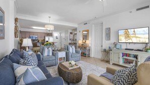 Ein geräumiges Wohnzimmer mit einer großen Couch, einem Couchtisch und einem Fernseher. Im Hintergrund befindet sich eine Küche mit Holzschränken und einem Essbereich.