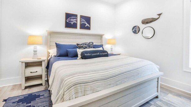 Un dormitorio con una cama grande, dos mesitas de noche y un tablero auxiliar. Hay dos cuadros enmarcados en la pared, un estante pequeño con objetos decorativos y una colcha a rayas azules y blancas.