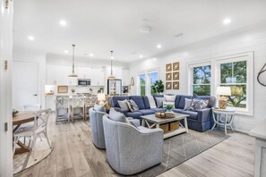 A spacious living room with a blue sofa set, a dining area with a wooden table, and a kitchen in the background. - Sweet Mama s 4 Bedrooms 4 Baths Cottage Beach Access Pool Sleeps 12 (Gulf Shores)