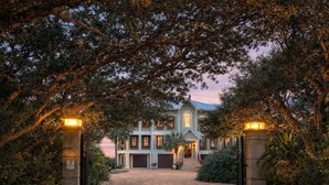 A large, two-story house with a driveway leading to a gated entrance, surrounded by trees and a clear sky. - The Gayosa House 9BR Beachfront Oasis With Private Pool Hot Tub (Orange Beach)