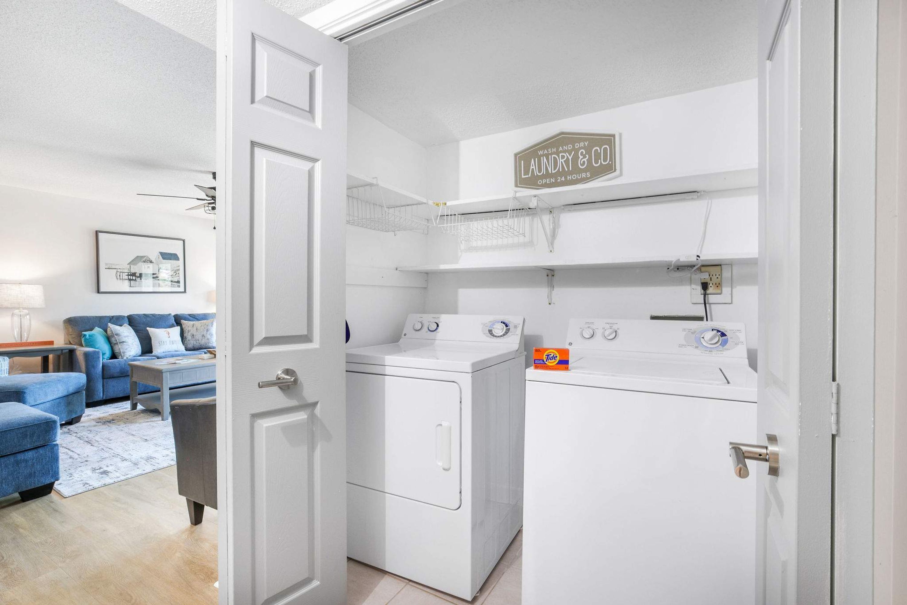 A laundry room with a washing machine and dryer, a shelf with laundry supplies, and a sign indicating 24-hour service.