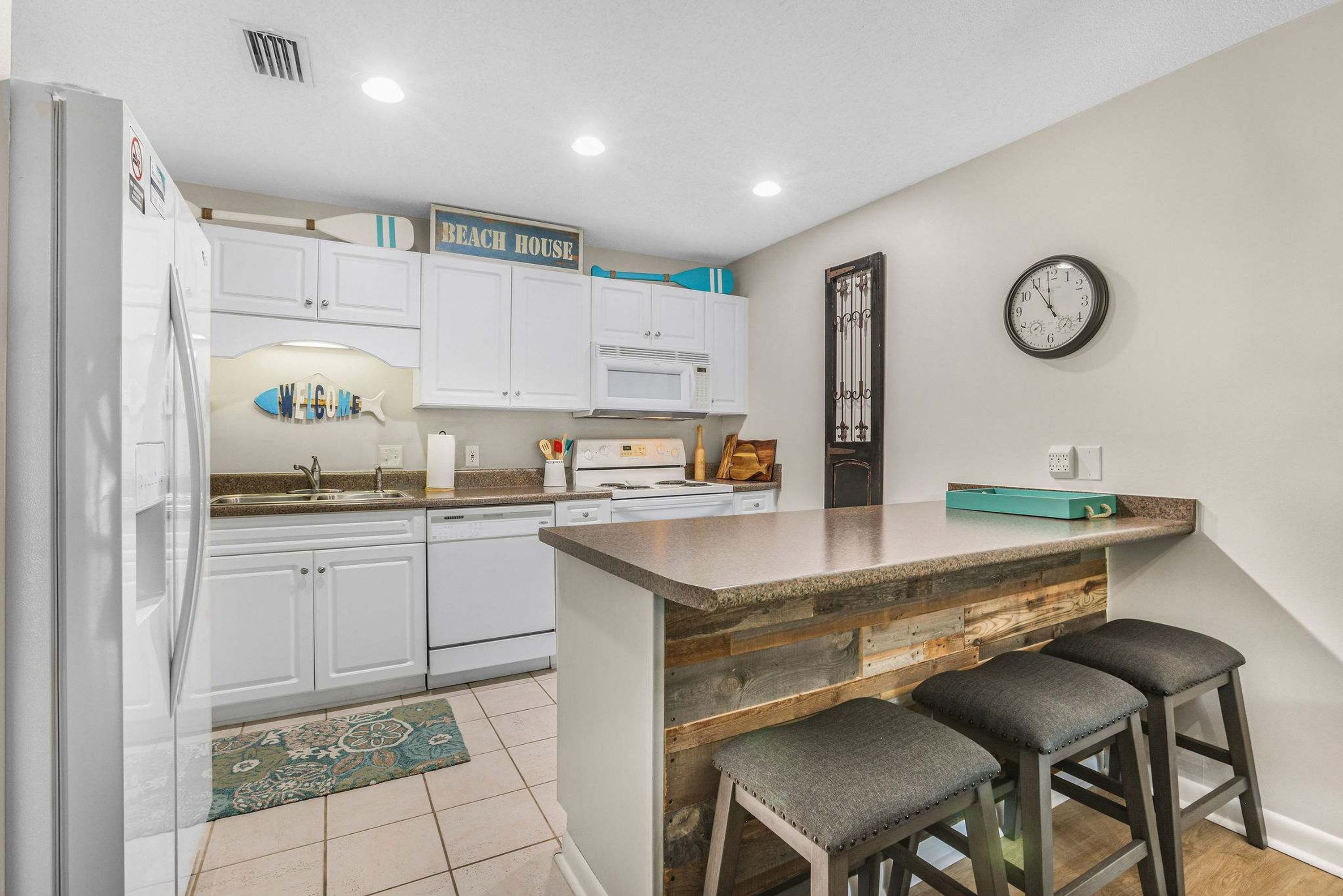 Une cuisine avec des meubles blancs, un îlot central en bois, une horloge et un panneau décoratif représentant une planche de surf.