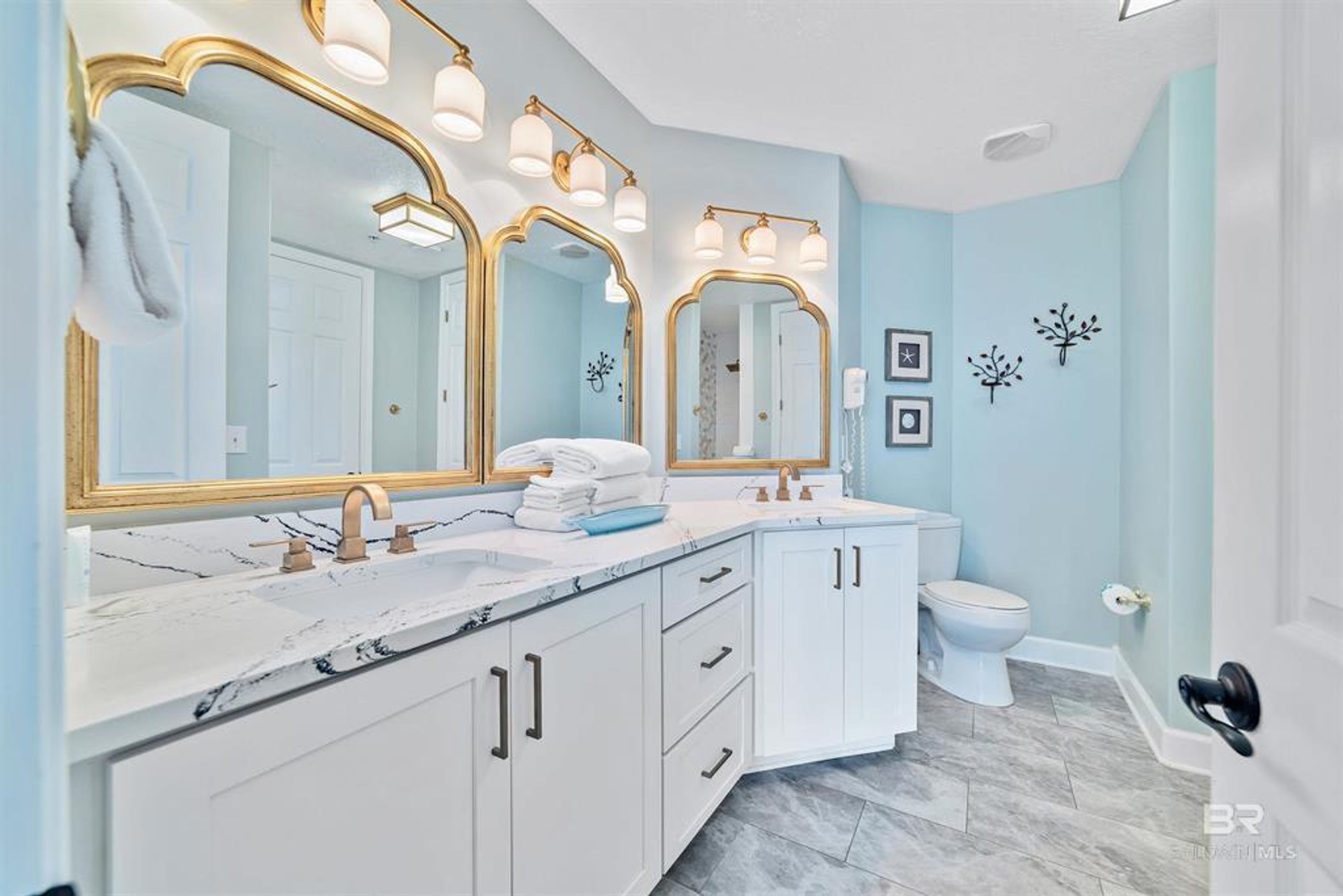 A bathroom with a white vanity, a double sink, a toilet, and a large mirror with gold frames.