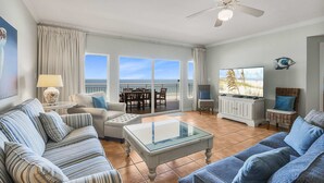 Een ruime woonkamer met een groot raam met uitzicht op de oceaan, een plafondventilator en een televisie op een witte kast.