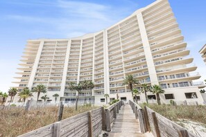 A multi-story hotel with a curved facade, a wooden boardwalk leading to the entrance, and palm trees in the foreground. - Admirals Quarters 508- Luxury Beachfront Condo 3 Bedrooms 3 Baths 5th Floor (Orange Beach)