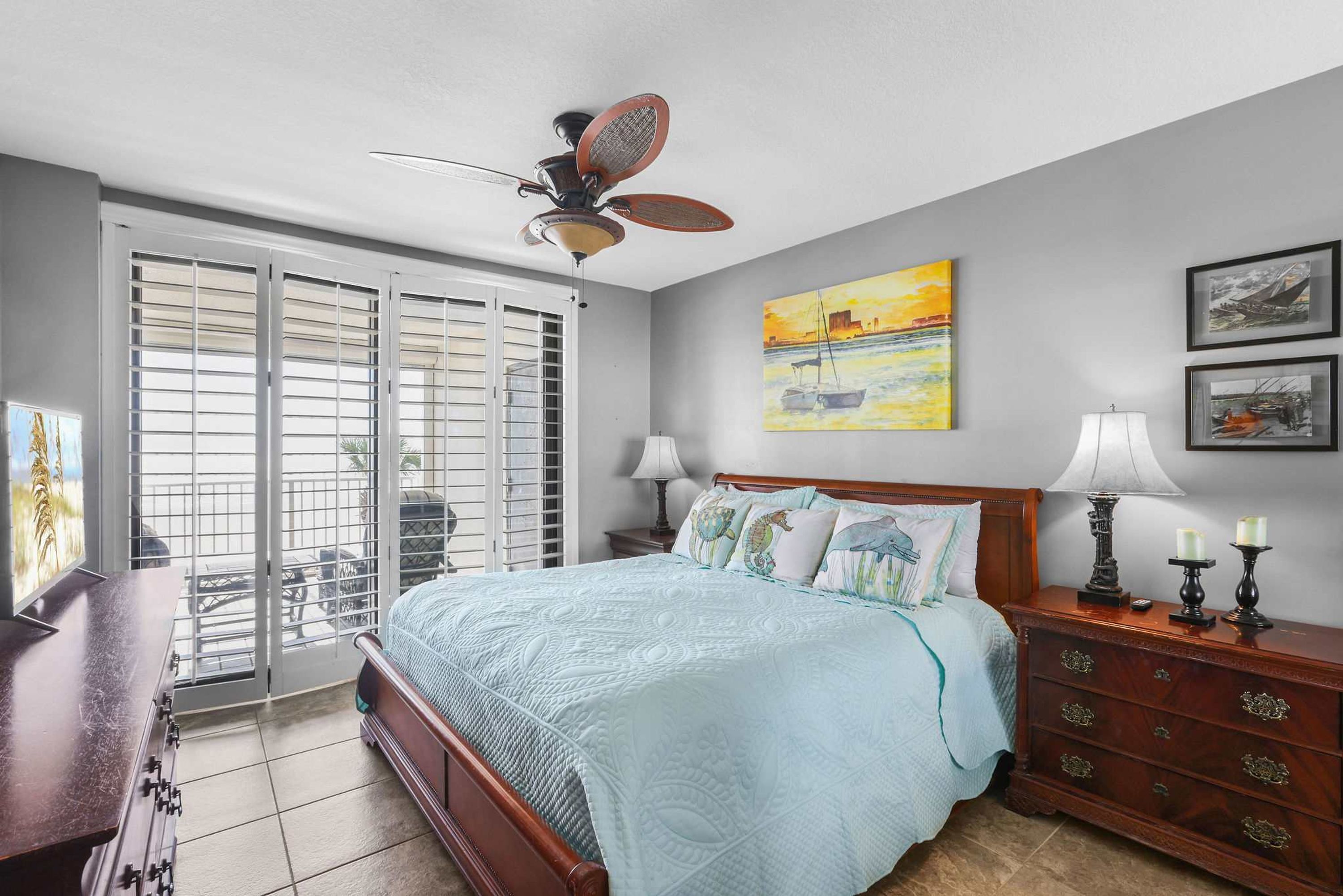 Une chambre à coucher comprenant un lit, une commode, un ventilateur de plafond, une fenêtre avec des stores et des œuvres d’art accrochées au mur.