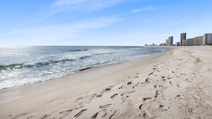 Ein sandiger Strand mit Fußspuren, die zum Wasser führen, ein klarer Himmel und eine Stadtansicht im Hintergrund.