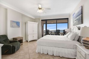 Ein Schlafzimmer mit einem großen Bett, einem Deckenventilator, Blick auf den Ozean, einem weißen Kleiderschrank und einem grünen Sessel.