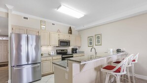 A kitchen with a stainless steel refrigerator, microwave, and stove, a bar area with white bar stools, and light-colored cabinetry. - Grand Pointe 310 Panoramic Views Huge Balcony 3rd Floor With 3 Bedrooms 3 5 Baths (Orange Beach)