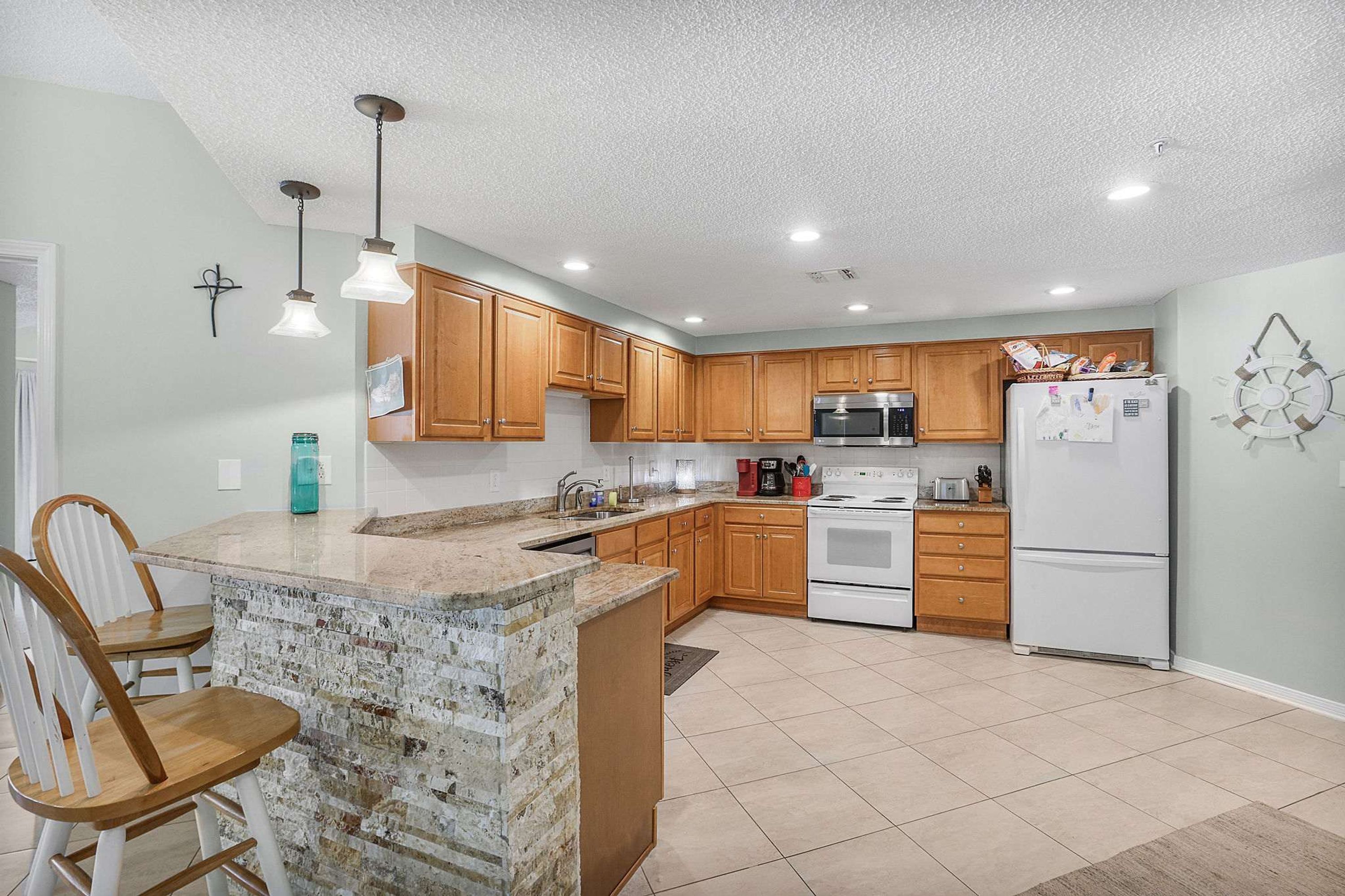 A kitchen with wooden cabinets, a granite island, a microwave, a refrigerator, and pendant lighting.