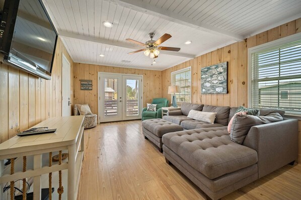 A cozy living room with a wooden interior, a ceiling fan, a television, and a large window with blinds. - Don t Worry Beach House 3 2 2 Blocks to Beach (Gulf Shores)