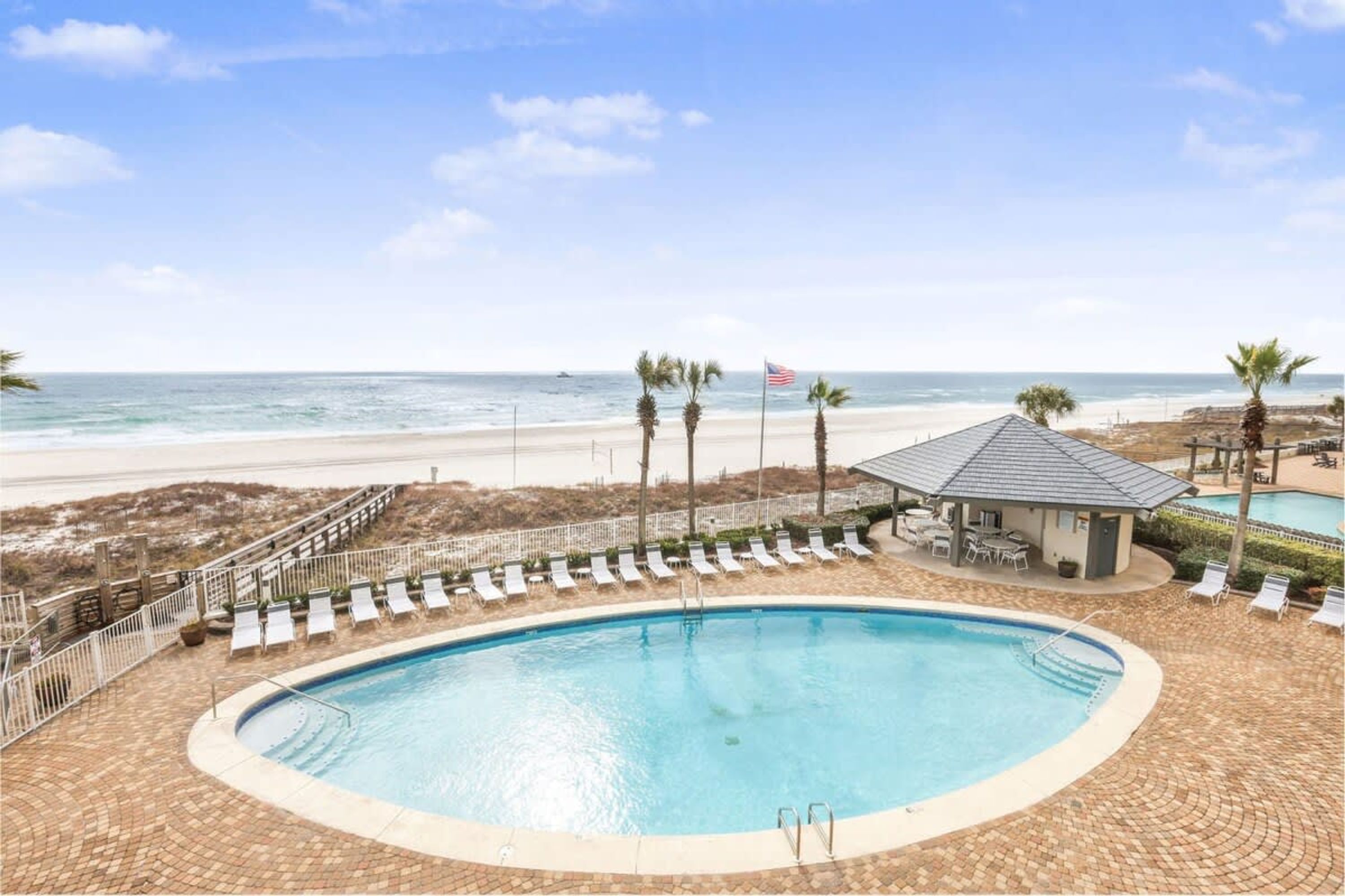 Zona de piscina con vista a la playa, sillas de descanso y un pequeño edificio.