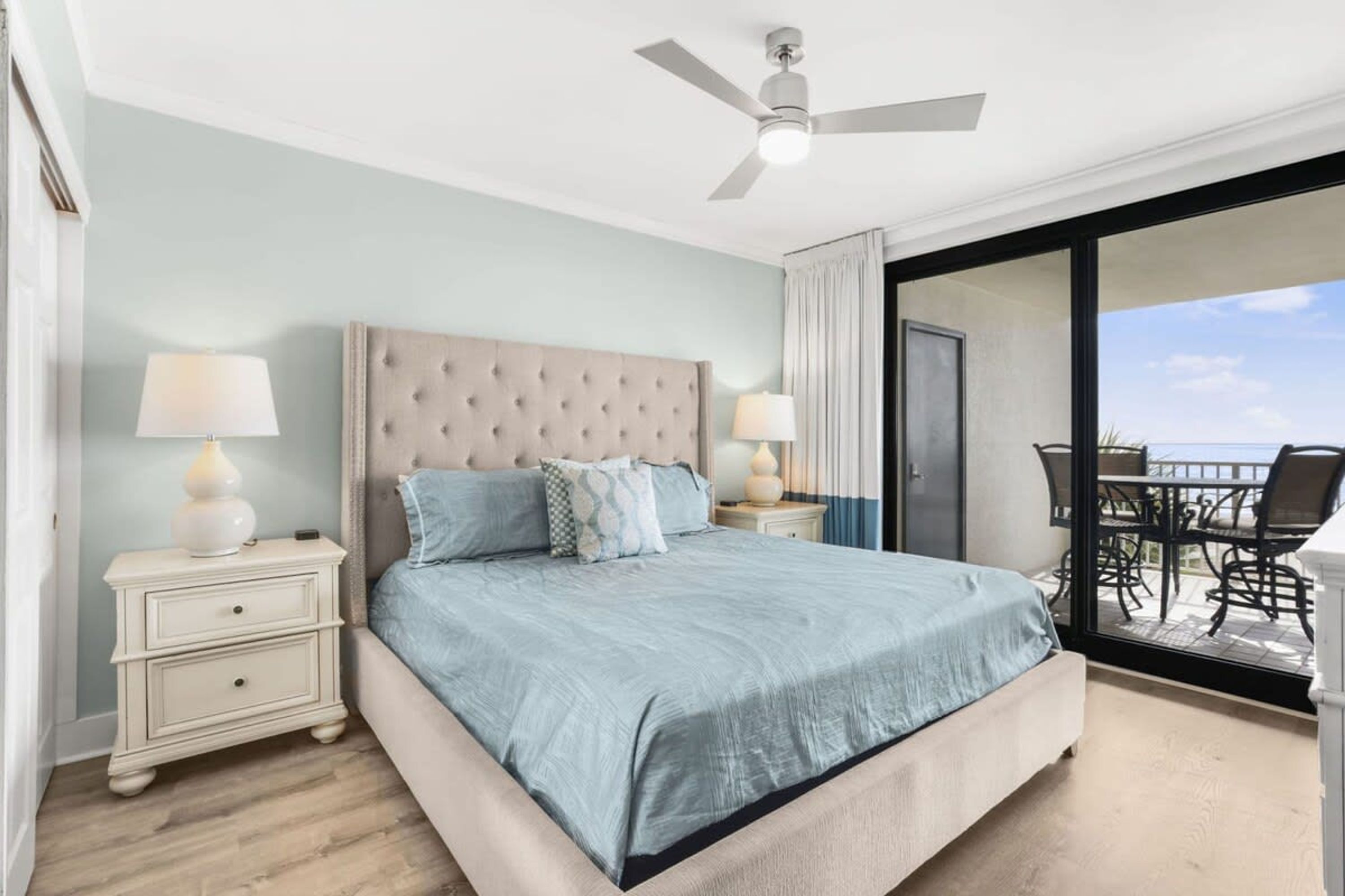 A bedroom with a large bed, two bedside tables with lamps, a ceiling fan, and a balcony with outdoor furniture.