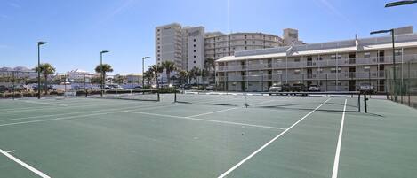 An outdoor tennis court with a net, surrounded by a fence and palm trees.