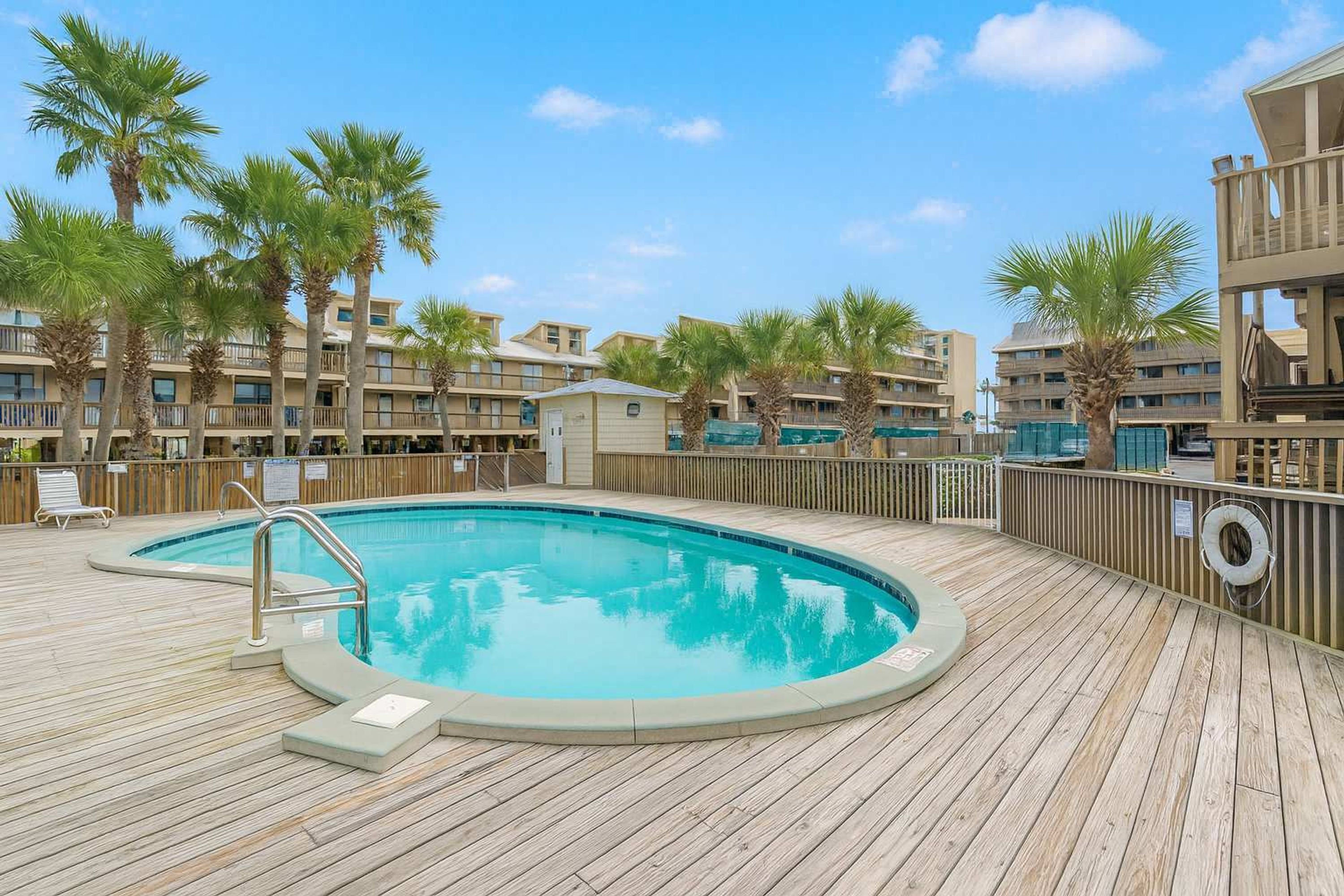 A wooden deck with a swimming pool, surrounded by palm trees and a multi-story building with balconies.