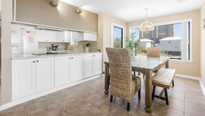 A modern kitchen with white cabinets, a dining area with a wooden table and woven chairs, and a large window with a view of buildings. - Compass Point 201 Pet-friendly for fee Boat Ramp (Gulf Shores)