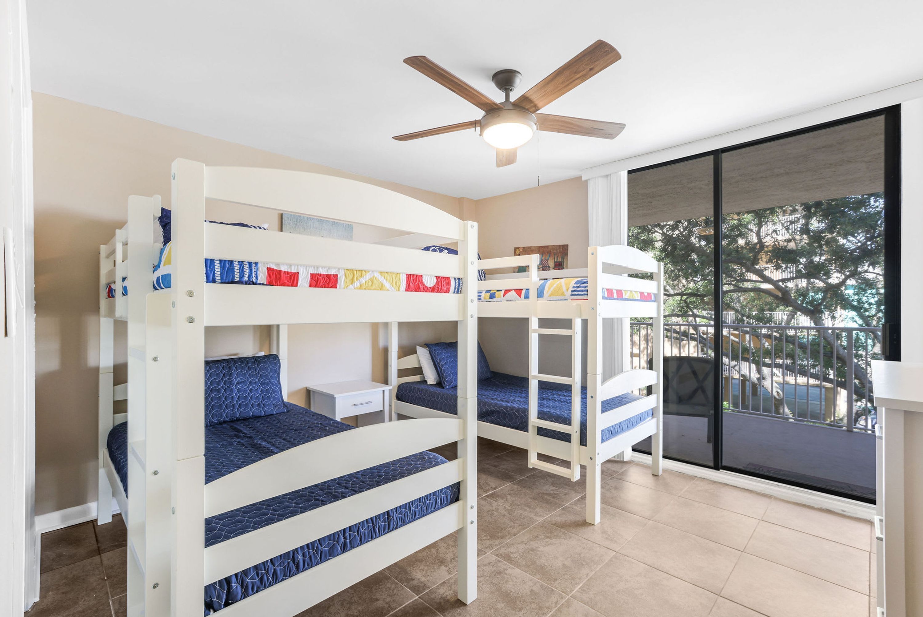 Une chambre avec un lit superposé, un ventilateur de plafond, une fenêtre donnant sur les arbres et un petit bureau.