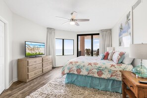 Ein Schlafzimmer mit Bett, Fernseher, Deckenventilator, Fenster und einer Tür, die auf einen Balkon führt.